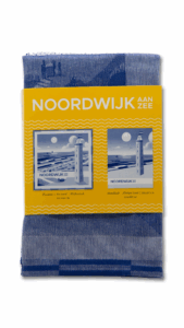 De blauw-witte keukendoek uit de Keukenset Noordwijk aan Zee-serie is gevouwen en voorzien van een geel label met een vuurtoren en meeuwen.
