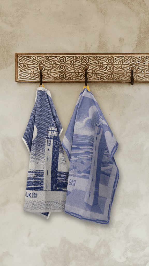De keukenset Noordwijk aan Zee bestaat uit twee blauw-witte theedoeken met een vuurtoren- en kustmotief, die aan een houten rek tegen een beige muur hangen.