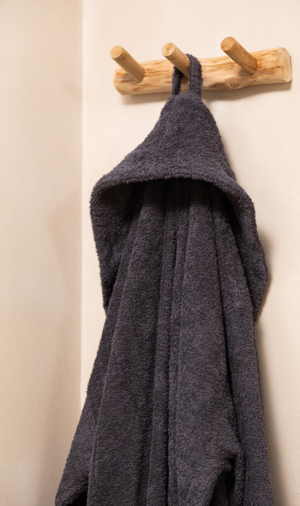 De Clarysse-badjas Billie in antraciet, in de maten large en extra large, hangt netjes aan een lichte houten wandhaak tegen een crèmekleurige muur.