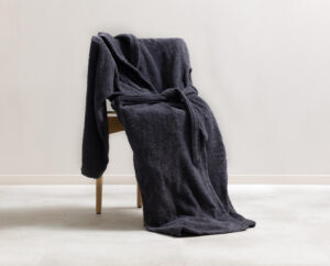 De Clarysse-badjas Billie Antraciet L XL is een zachte, donkergrijze badjas die over een houten stoel in een lichte kamer hangt.