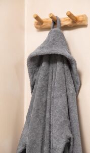 De Clarysse-badjas Billie Grafiet XXL is een grijze badjas met capuchon die aan een houten haak tegen een beige muur hangt.