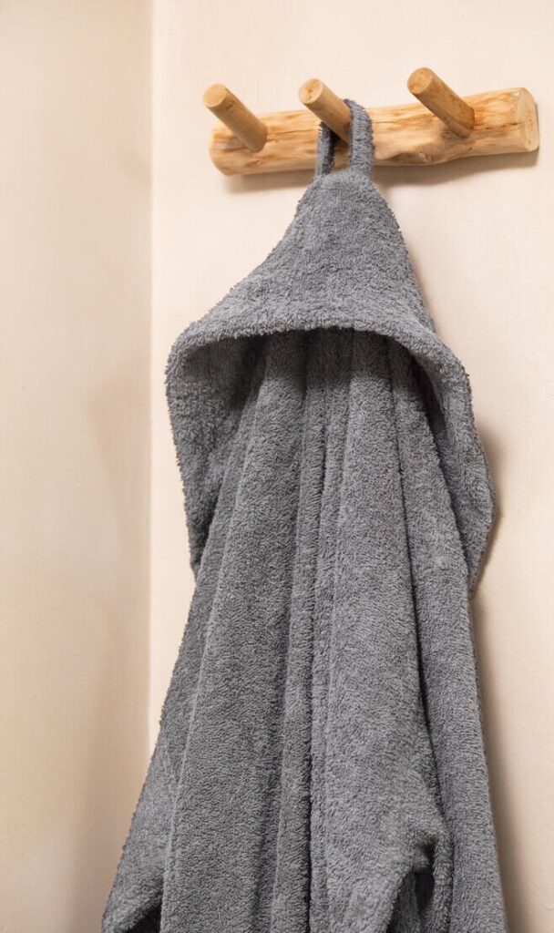 De Clarysse-badjas Billie Grafiet XXL is een grijze badjas met capuchon die aan een houten haak tegen een beige muur hangt.