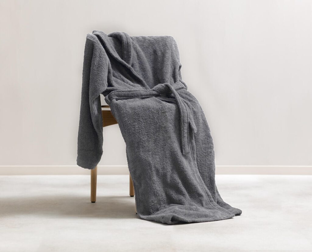 De Clarysse-badjas Billie Grafiet XXL, een zachte donkergrijze badjas met ceintuur, hangt over een houten stoel op een licht tapijt.