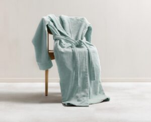 De Clarysse-badjas Flore in pastelgroen, maat ML, hangt nonchalant over een houten stoel, met losse mouwen en een losgeknoopte ceintuur.