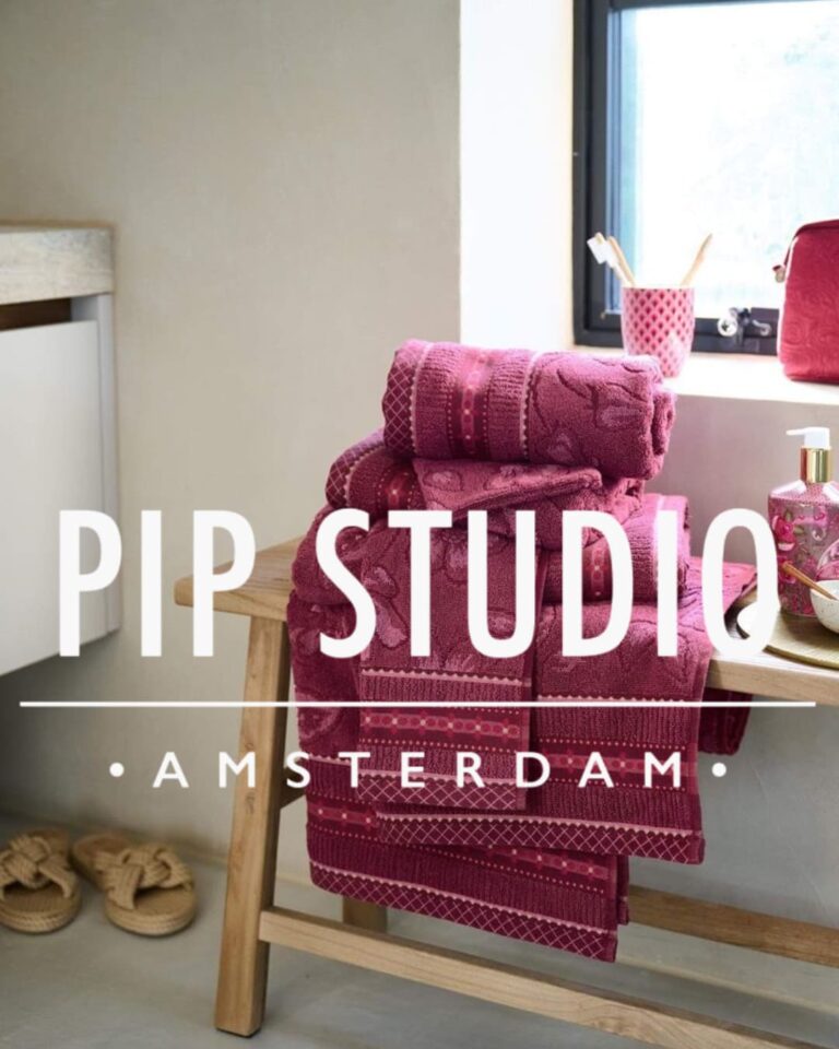 Op een houten bank bij het raam liggen donkerroze handdoeken met patroon van Merkaanbod Badgoed.com, aangevuld met accessoires van PIP Studio.