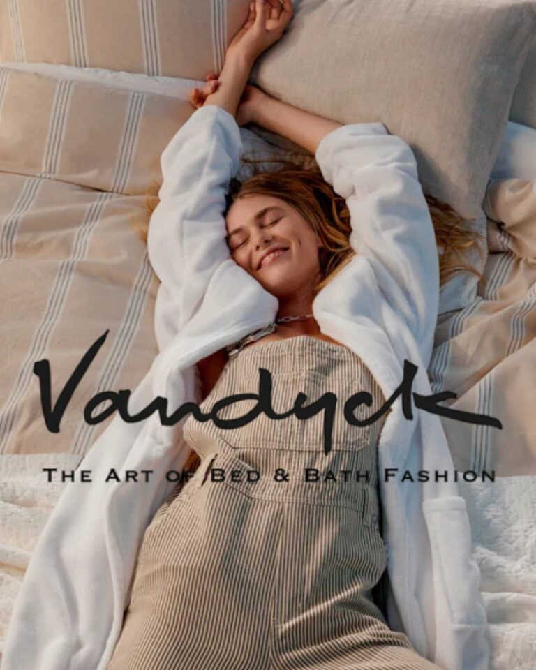 Een vrolijke vrouw in een witte badjas en een gestreepte overall strekt zich uit op een opgemaakt bed met beige en witte lakens van Vandyck uit de collectie van Merkaanbod Badgoed.com.