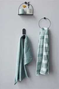 Twee groene Beddinghouse Sheer Handdoek-handdoeken hangen aan een grijze muur, met daarboven een plankje met bloemen en accessoires.