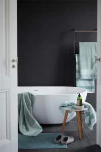 Een moderne badkamer met een vrijstaand bad, groene Beddinghouse Sheer Handdoek-handdoeken, een houten krukje met zeep en een flesje, grijze badslippers op de vloer en een donkere accentmuur.