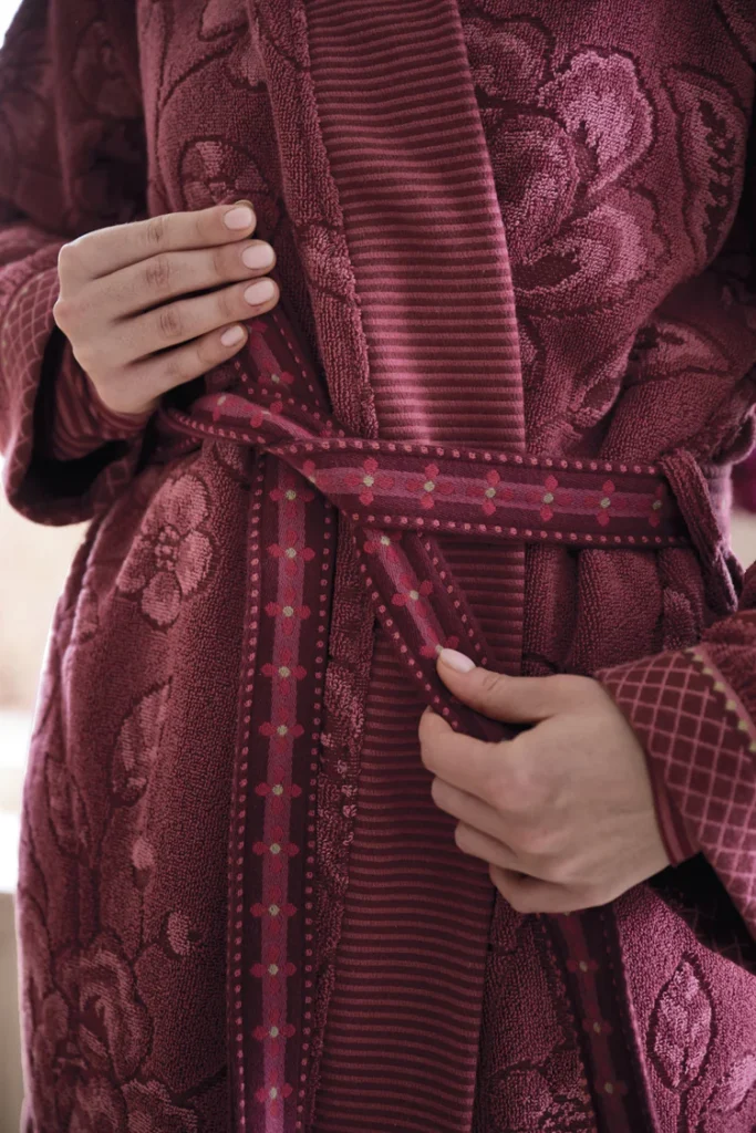 Close-up van iemand die de ceintuur van de Pip Studio Jasmin Jacquard-badjas (donkerroze, maat XL) om zijn of haar middel strikt, waarbij alleen de handen en het bovenlichaam in beeld zijn.