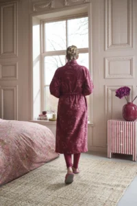Iemand draagt een Pip Studio Jasmin Jacquard-badjas in donkerroze, maat XXL, en pantoffels in een sfeervolle slaapkamer met roze beddengoed.