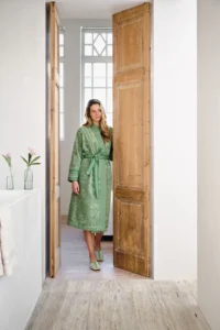 Een vrouw draagt een groene Pip Studio Jasmin Jacquard-badjas en staat in een deuropening met hoge houten deuren en roze bloemen.