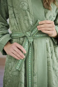Close-up van handen die de Pip Studio Jasmin Jacquard-badjas in groen (maat XL) vastknopen, met kleurrijke bloemen- en geometrische motieven.