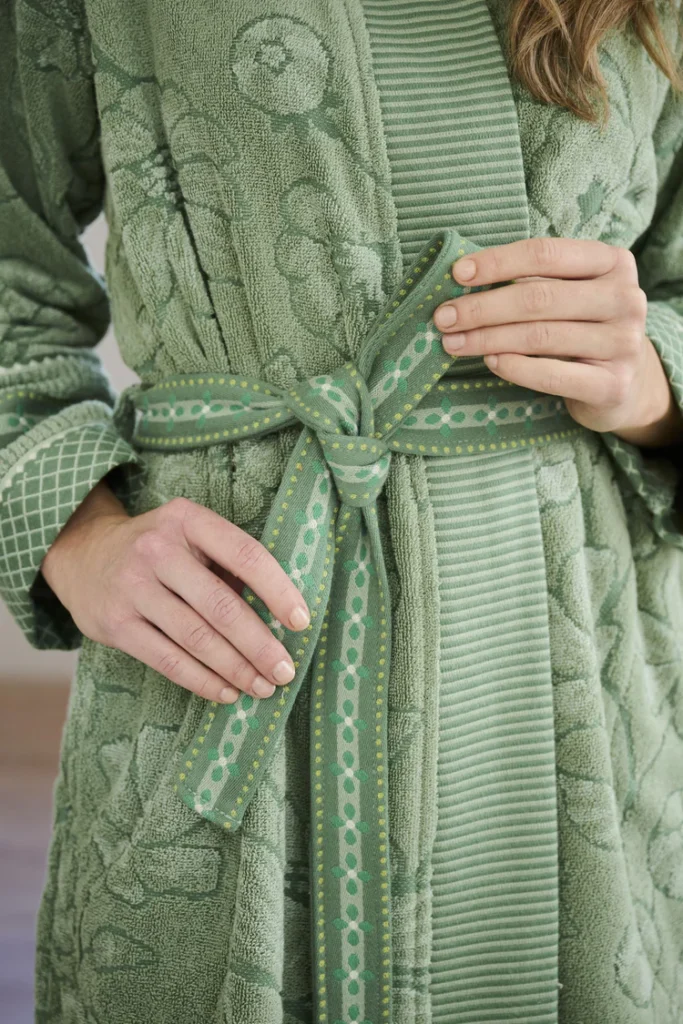Iemand knoopt de ceintuur van de Pip Studio Jasmin Jacquard-badjas in groen (XXL) vast; alleen zijn handen en een deel van zijn bovenlichaam zijn zichtbaar. De groene badjas heeft een subtiel reliëfpatroon, versierde manchetten en een bijpassende ceintuur.