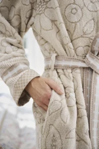 Een persoon draagt de Pip Studio Jasmin Jacquard-badjas in de kleur Sand, maat L, een beige badjas met bloemenpatroon en ceintuur, en heeft één hand in de zak.