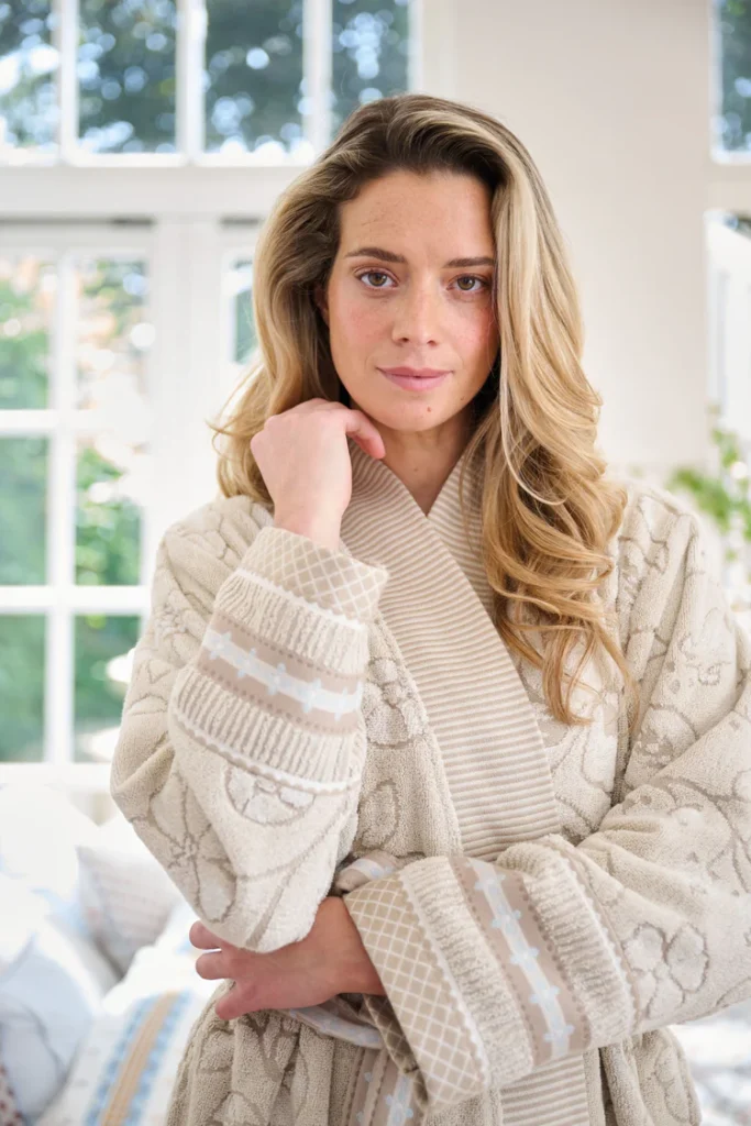 Een vrouw met lang blond haar poseert in de Pip Studio Jasmin Jacquard-badjas in de kleur Sand en kijkt zachtjes naar de camera.