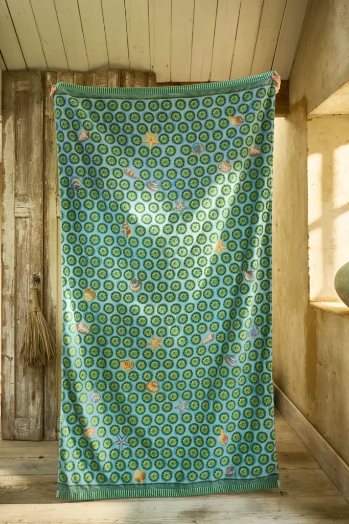 In een zonnige, rustieke kamer met houten vloeren en een houten deur ligt het Pip Studio Sea Dots-strandlaken in groen met blauwe cirkels en kleurrijke bloemenpatronen.