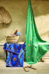Op een rustieke bank tegen een beige muur staan een rieten mandje met handdoeken, het Pip Studio Sea Dots-strandlaken in groen, geweven slippers, een blauwe handdoek met schelpenpatroon en een gedroogd palmblad.