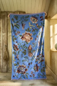 Het kleurrijke Pip Studio Secret Garden-strandlaken in blauw, met bloemen en vogels, ligt tegen een houten wand in een sfeervolle kamer.