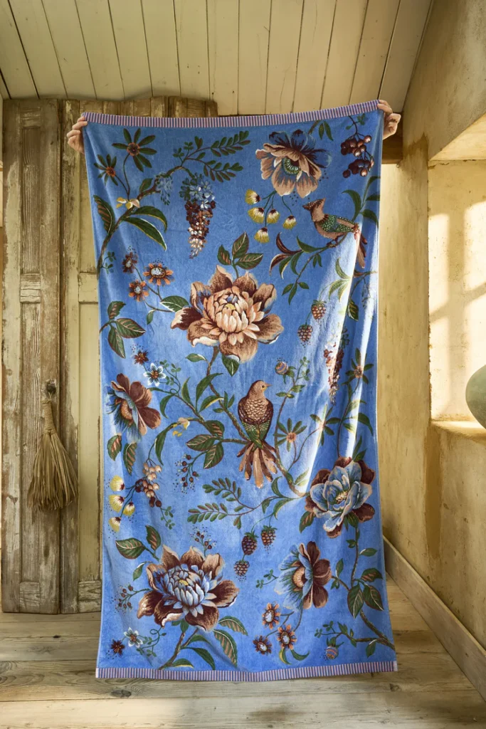 Het kleurrijke Pip Studio Secret Garden-strandlaken in blauw, met bloemen en vogels, ligt tegen een houten wand in een sfeervolle kamer.