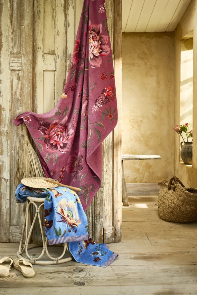 In een rustieke ruimte met houten wanden ligt het Pip Studio Secret Garden-strandlaken in roze tussen bloemen en zomerse accessoires.