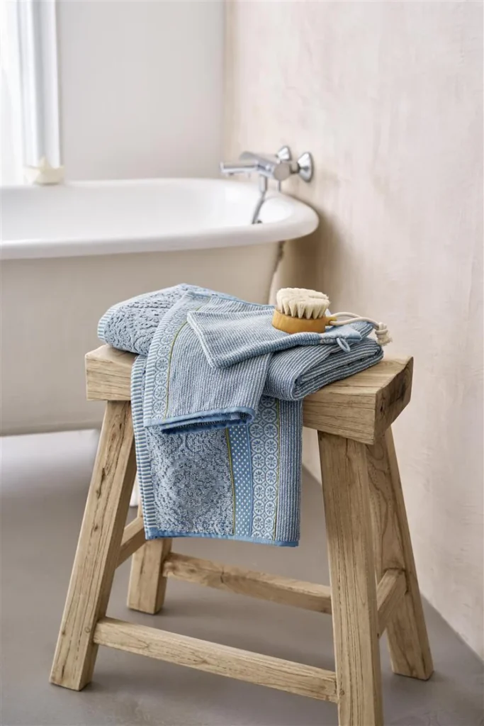 Op een houten kruk ligt de Pip Studio Soft Zellige-washandjeset in Blue Grey, voor een vrijstaand bad in sfeervol licht.