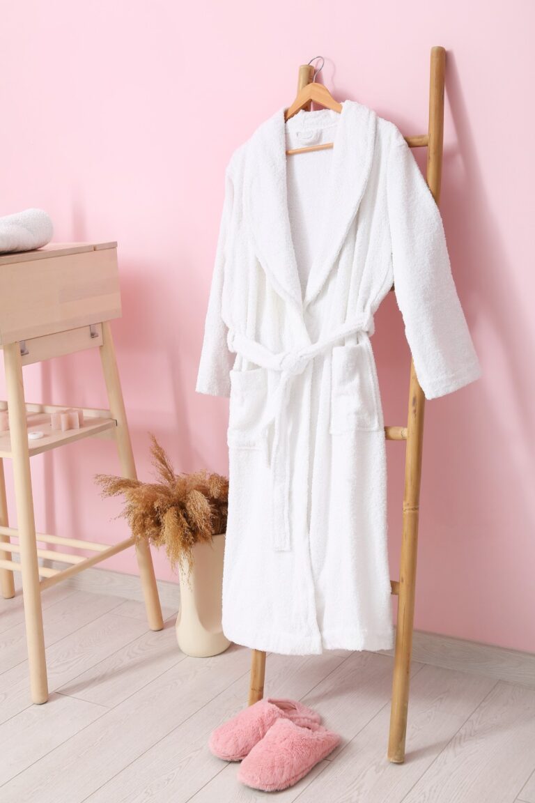 Een witte badjas hangt aan een houten ladder naast roze pantoffels, met een tafeltje en pampasgras in een vaas: een luxe moederdagcadeau.