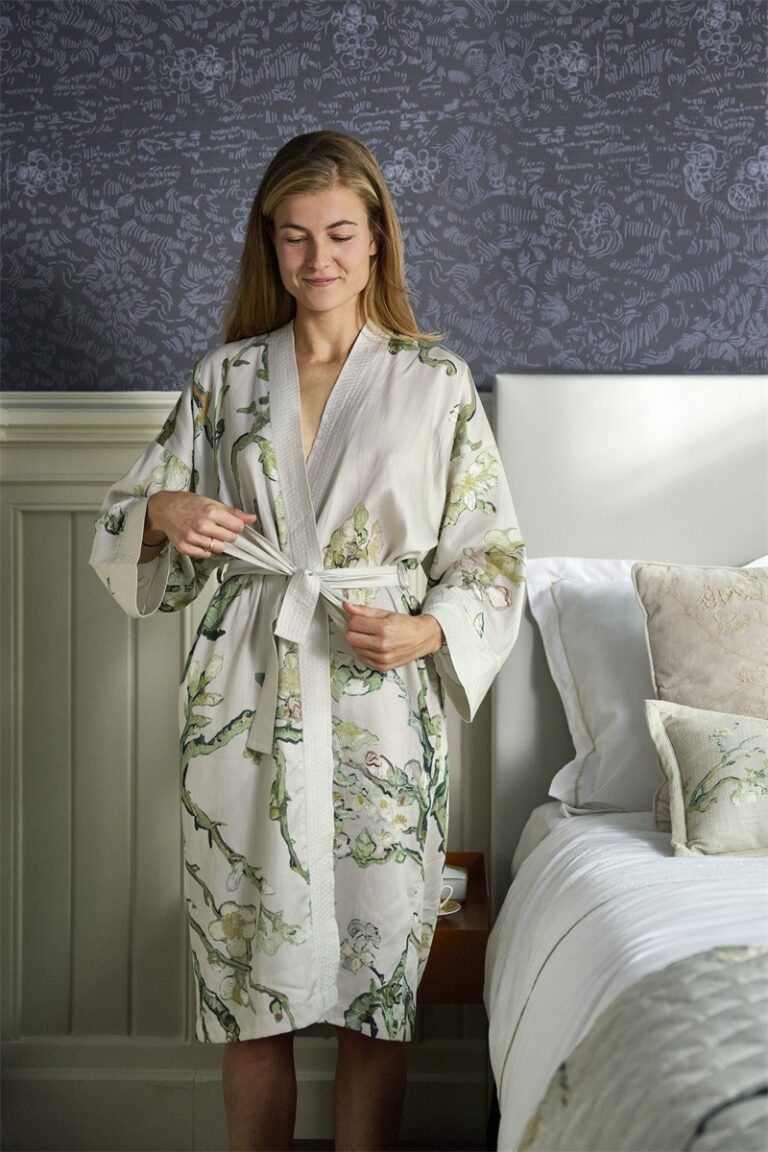 Een vrouw glimlacht zachtjes terwijl ze naast het bed de ceintuur van haar Van Gogh Almond Blossom-badjas in de kleur Sand (LXL) vastmaakt.