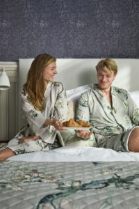 Een vrouw en een man, gekleed in de Van Gogh Almond Blossom-badjas in de kleur Sand, zitten lachend op een bed met een dekbed met botanisch motief in een kamer met donker bloemenbehang, terwijl de vrouw croissants vasthoudt.