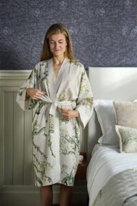 Een vrouw glimlacht terwijl ze de riem van de Van Gogh Almond Blossom Badjas Sand S/M vastmaakt, naast een opgemaakt bed met sierkussens.