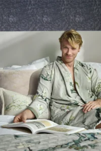Een man in een Van Gogh Blossom-badjas (groen, maat L) glimlacht terwijl hij ontspannen op bed zit en in een open boek bladert.