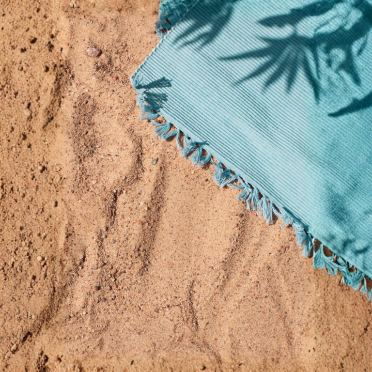 Op het zand ligt een lichtblauwe, gefranjerde handdoek van Badgoed.com, deels in de schaduw van palmbladeren.